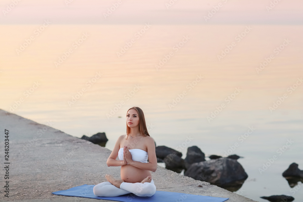 Young pregnant woman in white dress sitting on the beach near blue sea and breathing. Summer vacation during pregnancy, happy motherhood concept, close up on hands