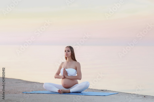 Young pregnant woman in white dress sitting on the beach near blue sea and breathing. Summer vacation during pregnancy  happy motherhood concept  close up on hands