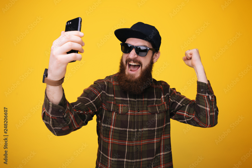 Happy excited young bearded man using cell phone and celebrating success over yellow background Happy excited young bearded man using cell phone and celebrating success over yellow background