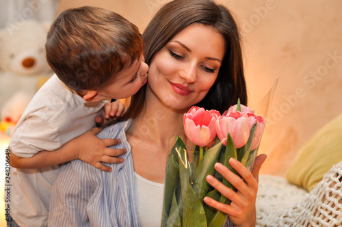 Little child son gives his mother a bouquet of delicate pink tulips and kisses her.