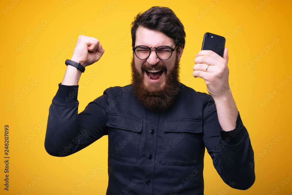 Portrait of a happy young bearded man in shirt holding mobile phone, celebrating victory, closed eyes Portrait of a happy young bearded man in shirt holding mobile phone, celebrating victory, closed eyes