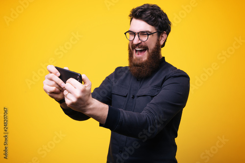 Image of excited young bearded man standing isolated over yellow wall background play games by mobile phone. © Vulp