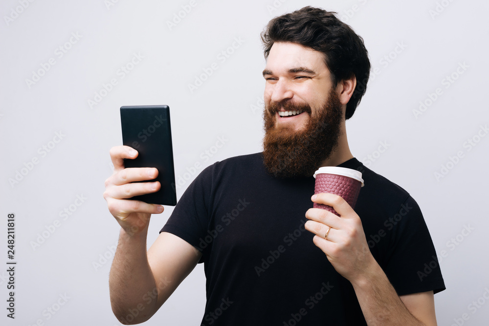 Excited bearded guy enjoying coffee break. Young man standing over white wall holding cup and using tablet. Excited bearded guy enjoying coffee break. Young man standing over white wall holding cup and using tablet.