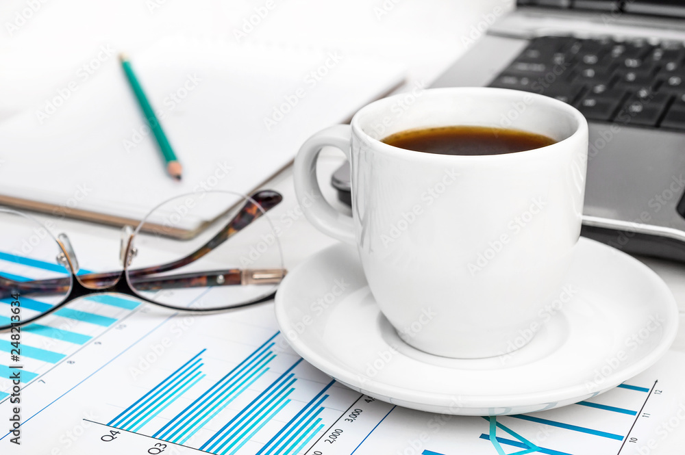 Cup of coffee with business graphs and laptop at office workplace. Cup of coffee with business graphs and laptop at office workplace.
