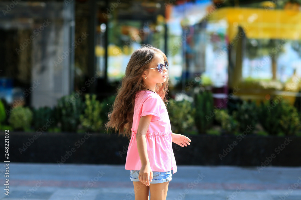 Naklejka premium Fashion kid summer in the city stands on a streetl. Trendy girl in sunglasses standing on the street at sunset.