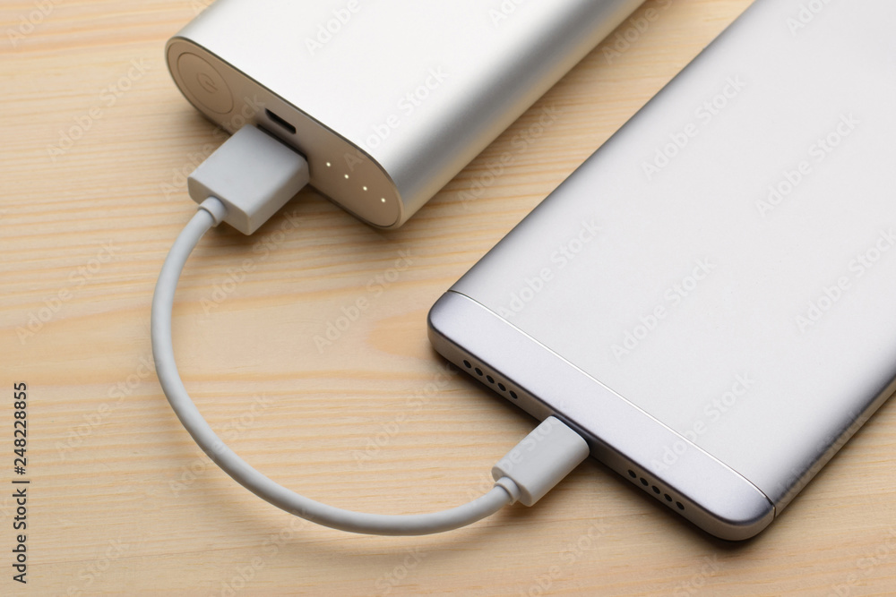 Silver smartphone charging with a power bank on a light wooden background Silver smartphone charging with a power bank on a light wooden background