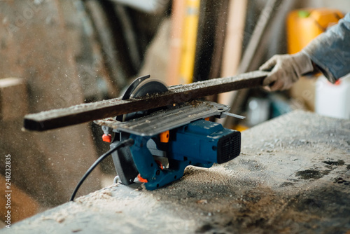 man in work clothes by profession carpenter handles wooden board on circular saw on a wooden workshop table, joiner works on machine in factory