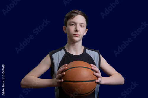 Portrait of a teen basketball player. The guy in the black t-shirt holds the ball in his hands. The concept of a sports poster or design news about basketball competitions. Isolated on blue background © romsvetnik