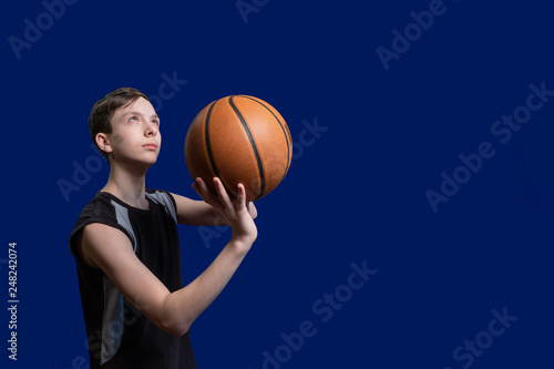 European teenager with a ball in his hands for playing basketball. The guy in the black shirt is preparing to throw the ball in the basket. Isolated on blue background. Copy space.