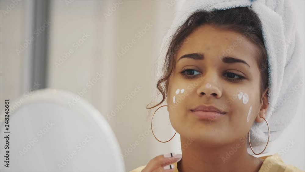 Beauty, skin care and people's concept - Smiling young woman. Applies a face cream and looking at the mirror at home. Beauty, skin care and people's concept - Smiling young woman. Applies a face cream and looking at the mirror at home.