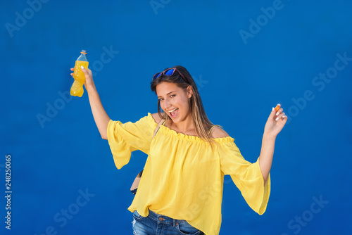 Cheerful pretty brunette in yellow blouse holding juice and laughing while standing in front of blue background.