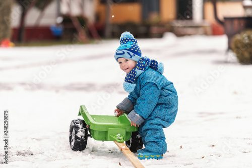 Happy toddler dressed in warm clothes playing with toy trailer on the snow. Holiday on country concept.