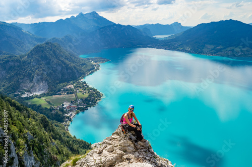 Exhausted but happy girl taking a break on a rock cliff above the turquise waters of Attersee lake, in Upper Austria, during a via ferrata route, on a bright, sunny, summer day.