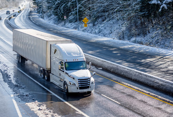 Big rig white bonnet semi truck with dry van semi trailer moving on the winding winter road with wet surface and snow
