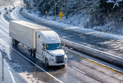 Big rig white bonnet semi truck with dry van semi trailer moving on the winding winter road with wet surface and snow photo