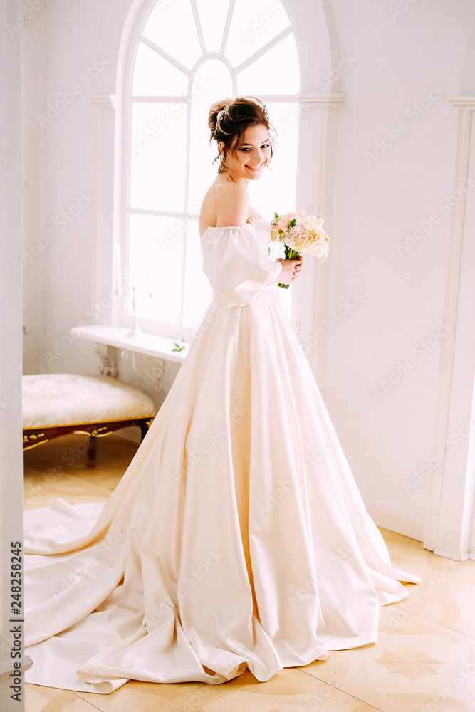 Elegant young woman in creamy satin dress standing near the window Elegant young woman in creamy satin dress standing near the window