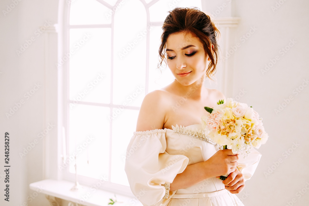 Elegant young woman in creamy satin dress standing near the window Elegant young woman in creamy satin dress standing near the window