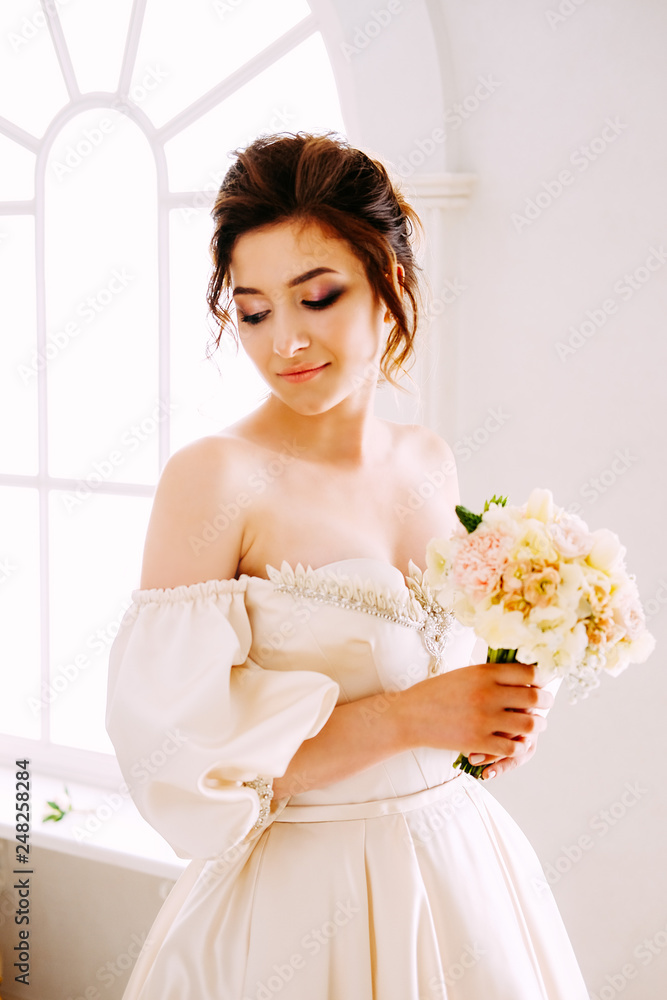 Elegant young woman in creamy satin dress standing near the window Elegant young woman in creamy satin dress standing near the window