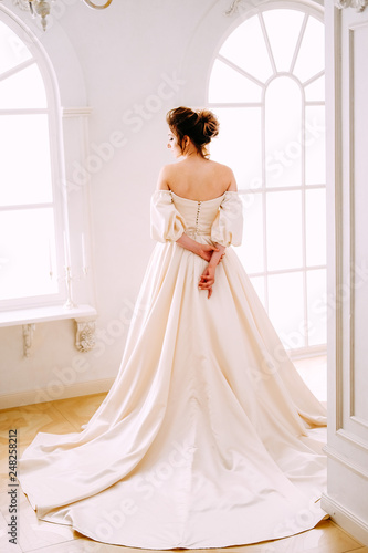 Elegant young woman in creamy satin dress standing near the window