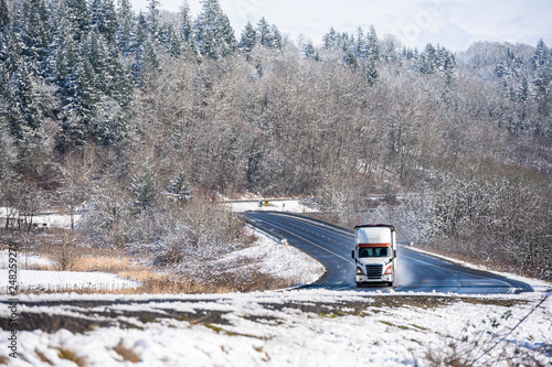 White and brown big rig semi truck transporting cargo in bulk semi trailer running on the winter snow winding road