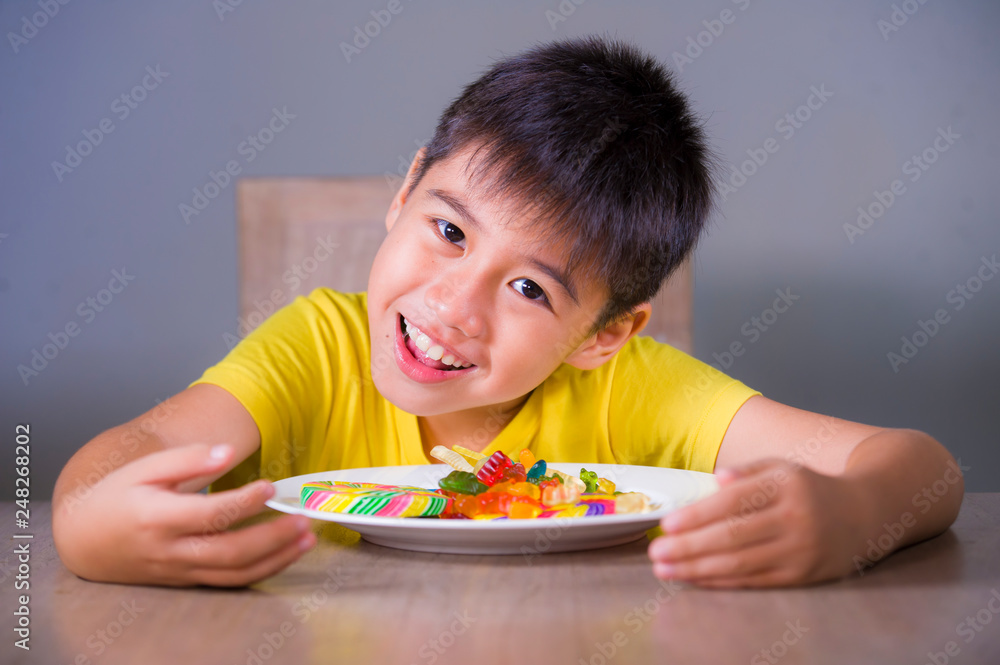 young happy and excited male kid smiling cheerful eating dish full of candy and lollipop sitting at table isolated on grey background in sugar addiction and abuse young happy and excited male kid smiling cheerful eating dish full of candy and lollipop sitting at table isolated on grey background in sugar addiction and abuse