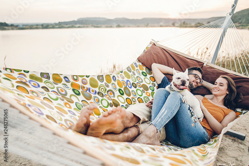 Hipster family on vacation concept, happy woman and man relaxing on a hammock at the beach with their cute bulldog pet, couple lying near a lake at sunset