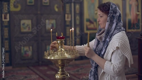 woman in the Russian Orthodox Church with red hair and a scarf on her head lights a candle and prays in front of the icon.