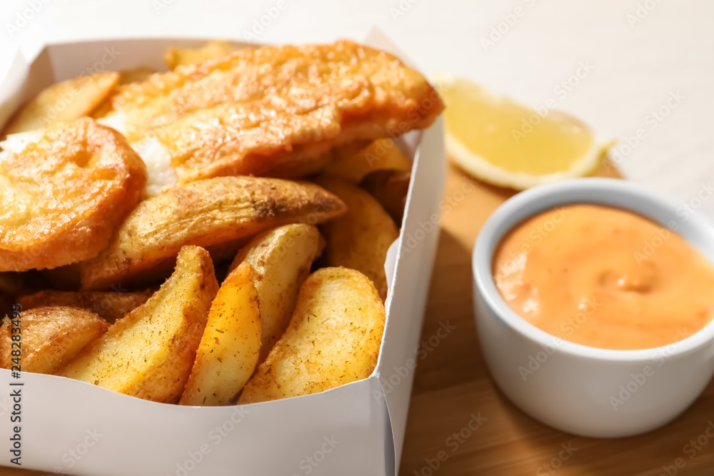 Disposable container with British Traditional Fish and potato chips on wooden board, closeup Disposable container with British Traditional Fish and potato chips on wooden board, closeup