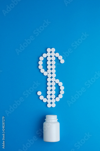 Multiple white tablets of drugs or vitamins on reflective grey surface spelling dollar sign to indicate high cost of medication or drugs. Vertical arrangement.