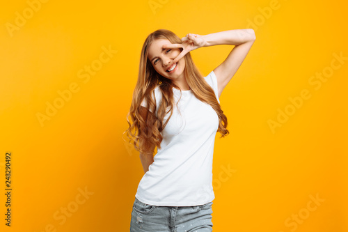 Portrait of a cheerful girl showing two fingers with winking eyes showing tongue against yellow background