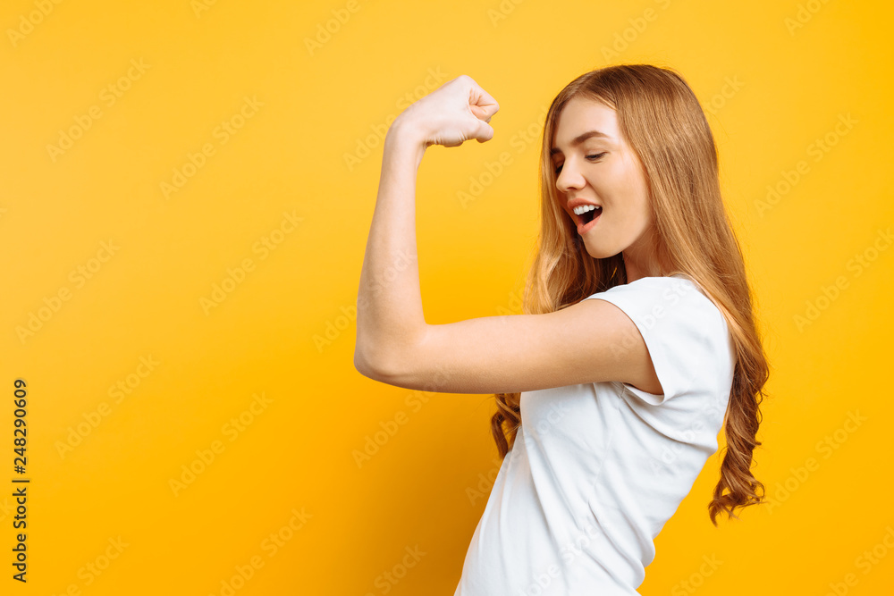 Φωτογραφία of happy girl in white t-shirt showing arm muscles on yellow background