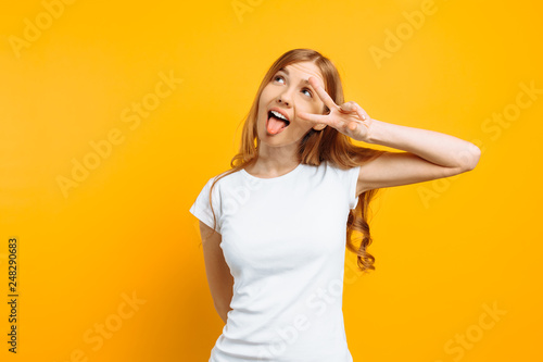 Portrait of a cheerful girl showing two fingers with winking eyes showing tongue against yellow background