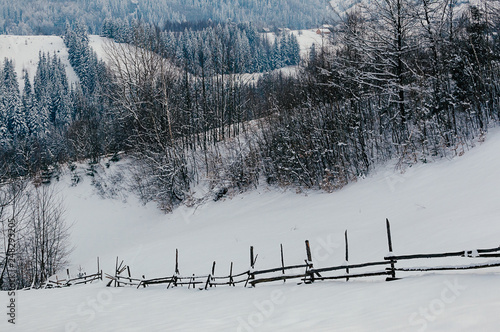 Wooden fence covered with snow cap. Winter rural landscape, village or farm, snow-capped mountains and forest in the background © Kalinova Olena