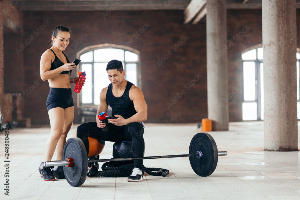 Obraz premium young woman and man with smartphone and water resting at gym. full length photo. copy space.online connection