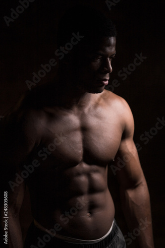 Handsome African American bodybuilder man, with naked muscular torso posing against red brick wall. Low key photo.