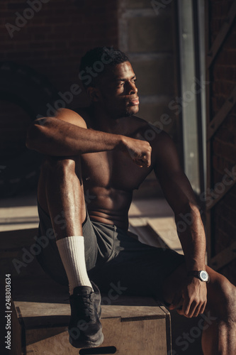 Young afroamerican athletic man with naked perfect shape muscular torso sitting isolated over brick background.
