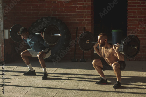 Couple of shirtless interracial male athletes pumping muscles with heavy weight barbell at indoor crossfit studio