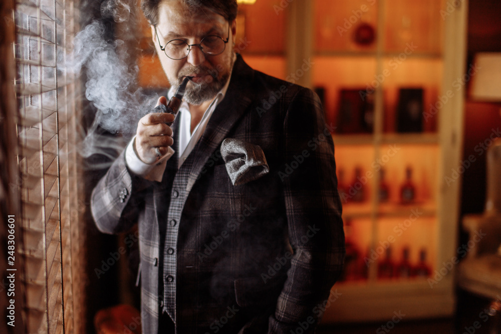 Portrait of confident mature businessman in expensive luxury english retro  style suit smoking tobacco pipe over dark living room interior with wine  shelves on background Stock Photo | Adobe Stock