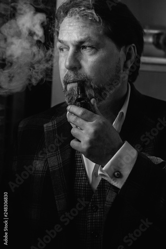 White and black portrait of well-dressed mature bearded caucasian man, small business owner, in expensive tailored suit relaxing with tobacco pipe