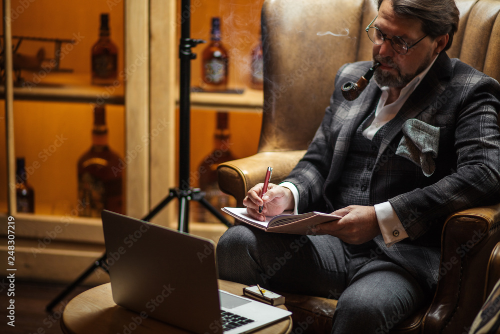 Handsome mid-age male blogger wearing trendy tweed jacket, puffing tobacco pipe and working on cafe table with laptop computer, writing new article in his blog. Handsome mid-age male blogger wearing trendy tweed jacket, puffing tobacco pipe and working on cafe table with laptop computer, writing new article in his blog.