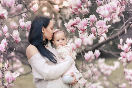 Cute Baby 6 month old Girl in Pink Outfit with Big Blue Eyes with Young Beautiful Mother at Spring, Pink Blooming Tree at the Background