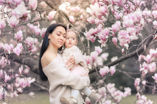 Cute Baby 6 month old Girl in Pink Outfit with Big Blue Eyes with Young Beautiful Mother at Spring  Pink Blooming Tree at the Background