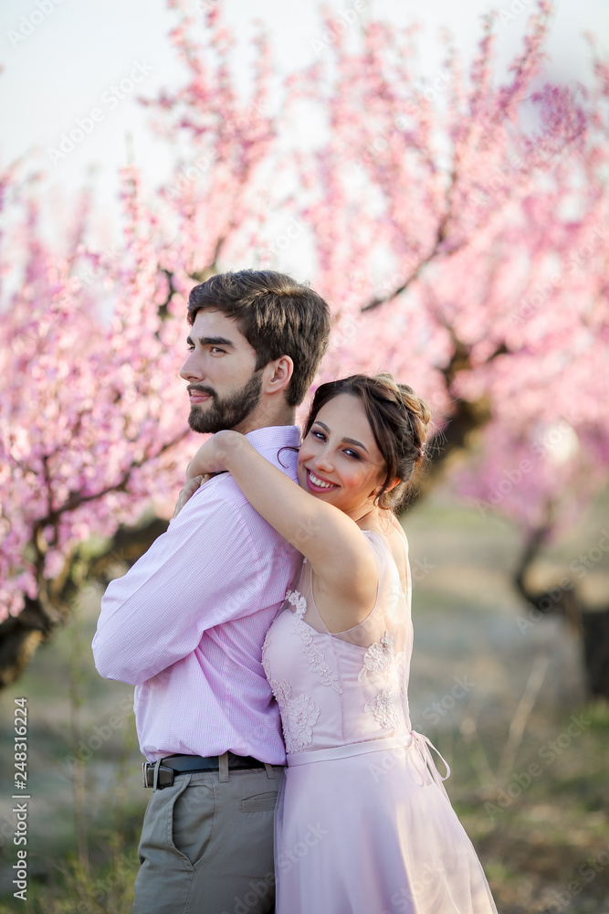 Newlyweds in love stand on the nature, against the background of wooden stakes, in sunny weather. Stylish groom embraces a beautiful bride in a lace dress in a green garden. Newlyweds in love stand on the nature, against the background of wooden stakes, in sunny weather. Stylish groom embraces a beautiful bride in a lace dress in a green garden.