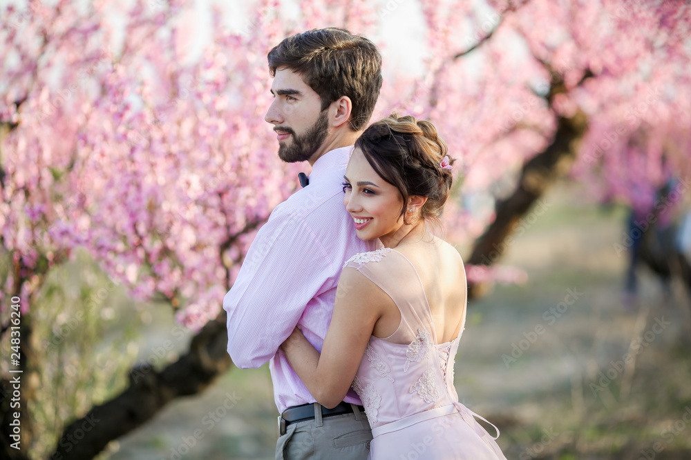 Newlyweds in love stand on the nature, against the background of wooden stakes, in sunny weather. Stylish groom embraces a beautiful bride in a lace dress in a green garden. Newlyweds in love stand on the nature, against the background of wooden stakes, in sunny weather. Stylish groom embraces a beautiful bride in a lace dress in a green garden.