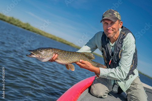 Happy fisherman with big pike fish trophy at the boat with fishing tackles