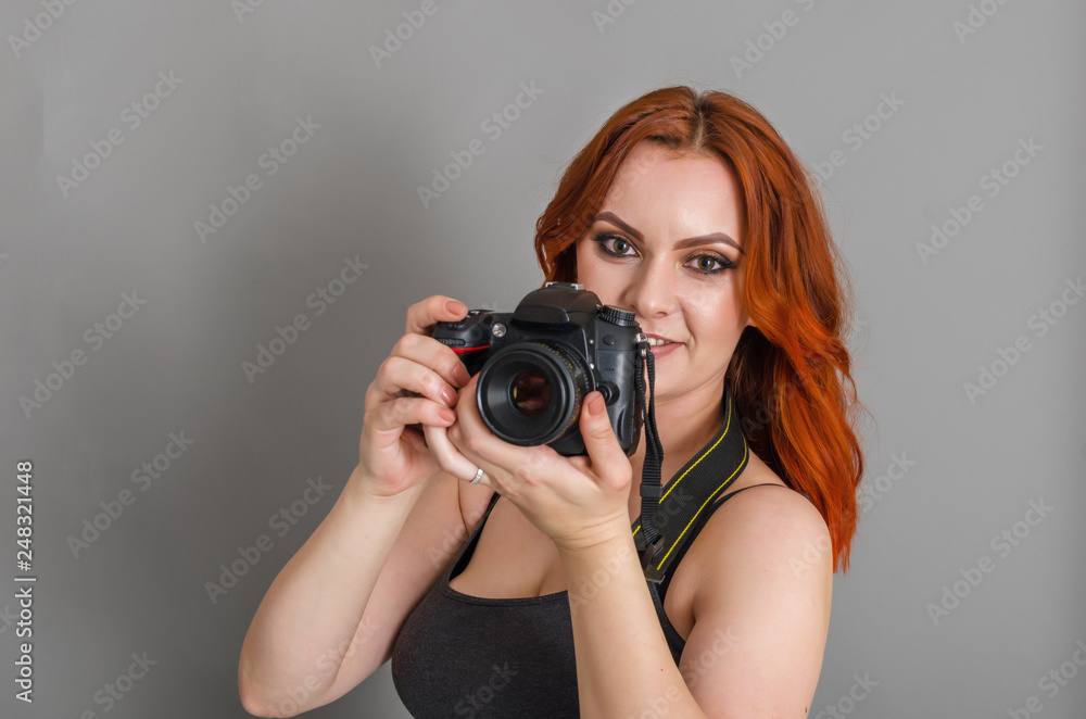Smiling woman with red hair holds a modern camera in her hands. Gray background, creativity, photographer, beautiful Smiling woman with red hair holds a modern camera in her hands. Gray background, creativity, photographer, beautiful