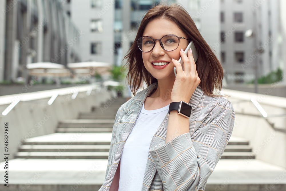 Business communication. Young lady in eyeglasses standing on the city street talking on smartphone joyful close-up