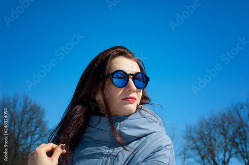Portrait of beautiful young woman with mirrored sunglasses outdoors over blue sky during early spring time.