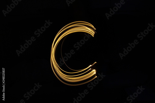 Long exposure, light painting photography. Letter c in a vibrant neon metallic yellow gold colour against a black background. Alphabet series.