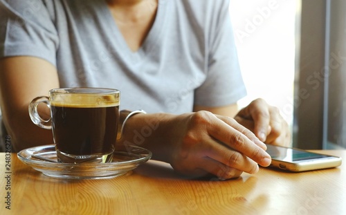 Slika na platnu The hand of the woman who was scrolling for information on the screen on her mobile phone  and hot coffee in clear glass that she had prepared for drinking